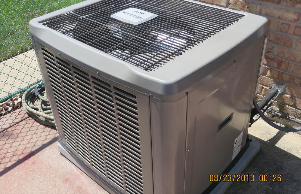 The outdoor condenser unit of a central air conditioning system beside a brick house, its aluminum fins visible through the side louvres