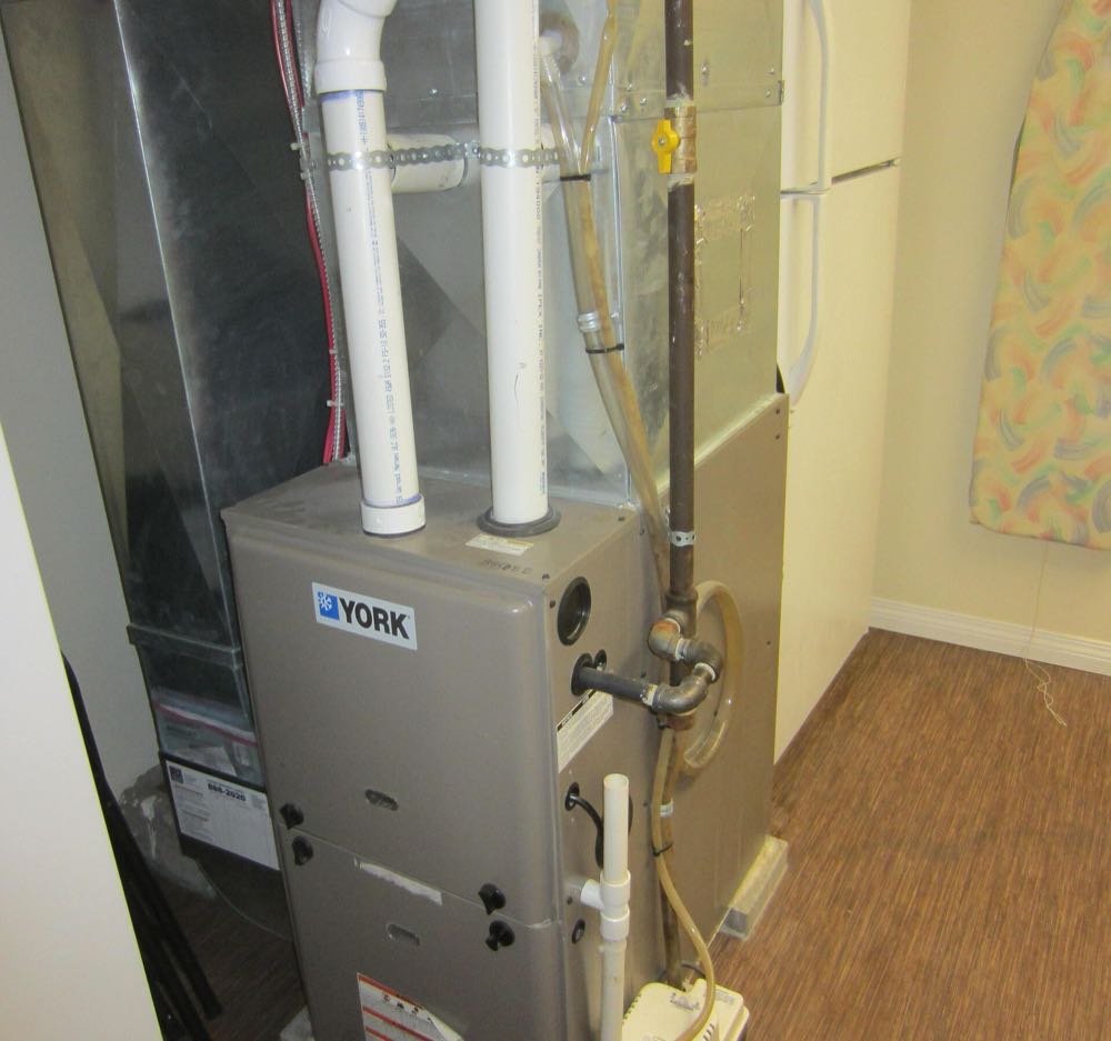 A newly installed high-efficiency condensing gas furnace with white PVC intake and exhaust pipes and a condensate pump beside it