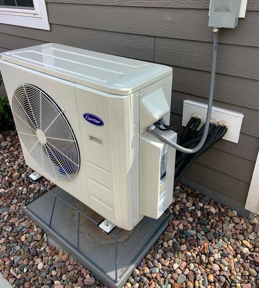 The outdoor unit of a residential air-source heat pump mounted on a stand beside a house, with the electrical disconnect box on the wall above it and insulated refrigerant lines running inside